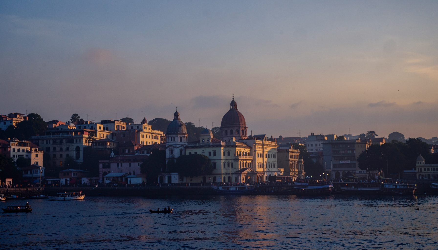 Warm golden-hour view over a riverside city skyline with domed buildings and boats, glowing amber and navy tones evoking Kolkata at dusk