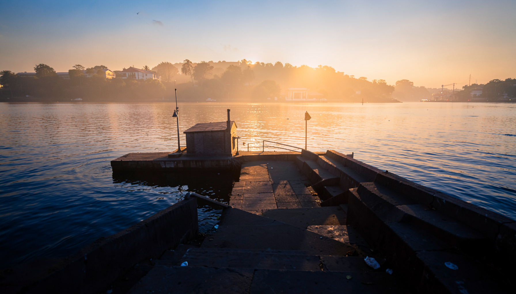 Warm golden-hour photo of a quiet riverside ghat with steps leading to the water in soft amber light